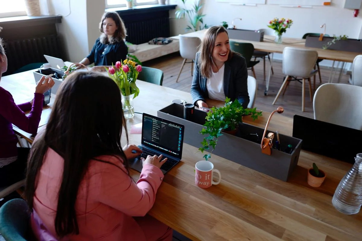 Group of people working together in a bright, modern co-working space with laptops, plants, and a casual, collaborative atmosphere.