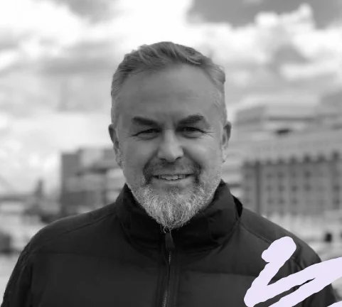 An image of AIM-FOR founder Lee Richardson smiling wearing a black jacket, with a blurred cityscape in the background, presented in black and white.