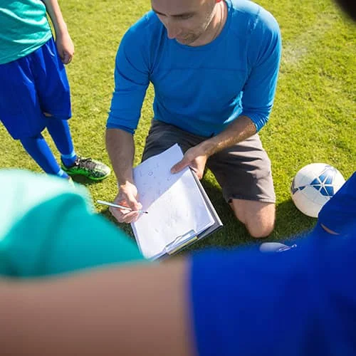 A football coach in a blue shirt kneeling on the grass, demonstrating tactics on a clipboard to players in blue kits during a training session.